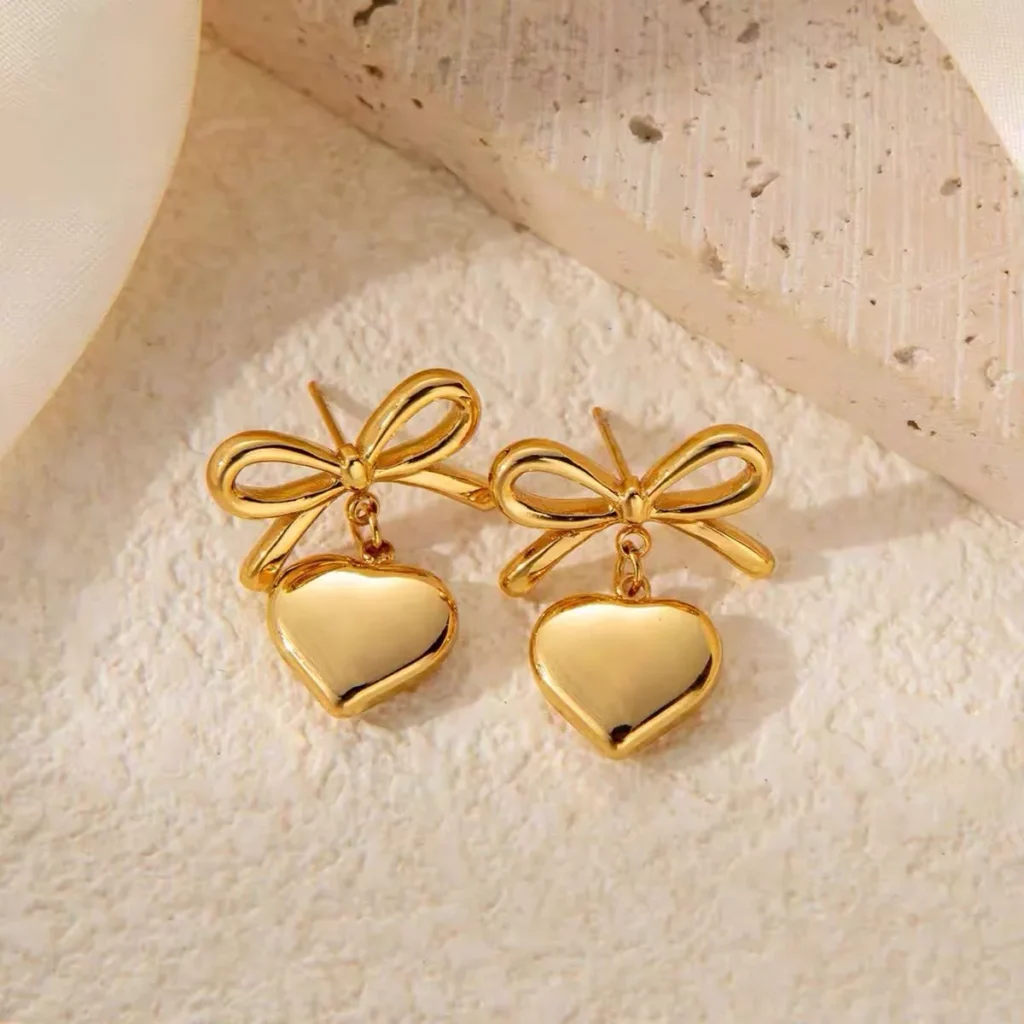 A pair of polished gold stainless steel earrings featuring heart-shaped drops and delicate bow designs, displayed on a textured beige background – ideal for wholesale fashion jewelry promotion.