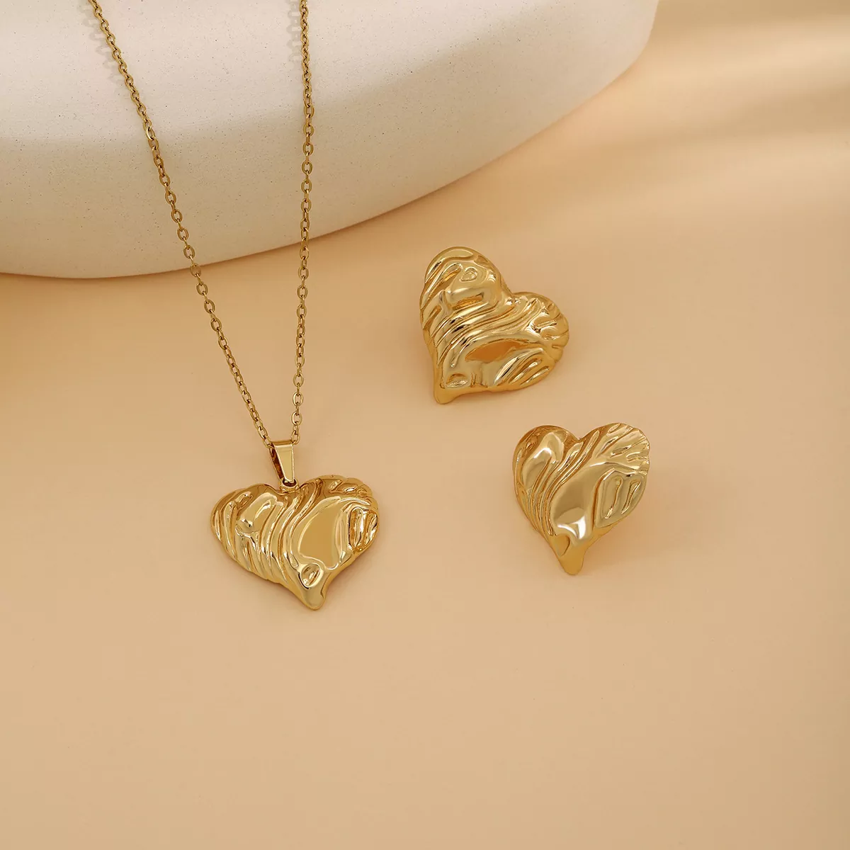 A digital photograph features a gold stainless steel jewelry set including a heart-shaped pendant necklace and matching earrings, displayed on a soft beige background.