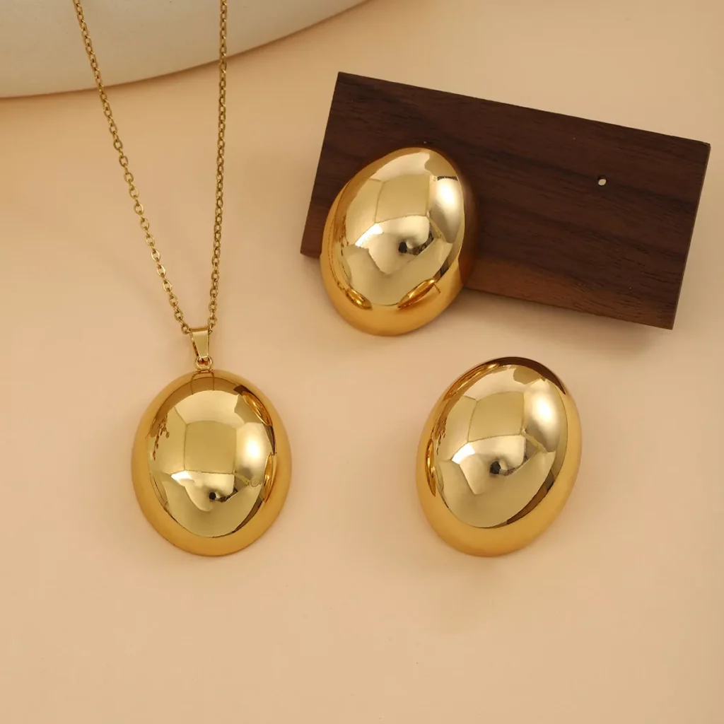 Glossy oval stainless steel jewelry set including a gold-tone pendant necklace and matching earrings, ideal for wholesale suppliers and resellers.