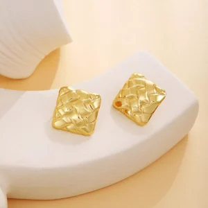 Gold square woven texture stainless steel earrings displayed on a white ceramic stand with warm background