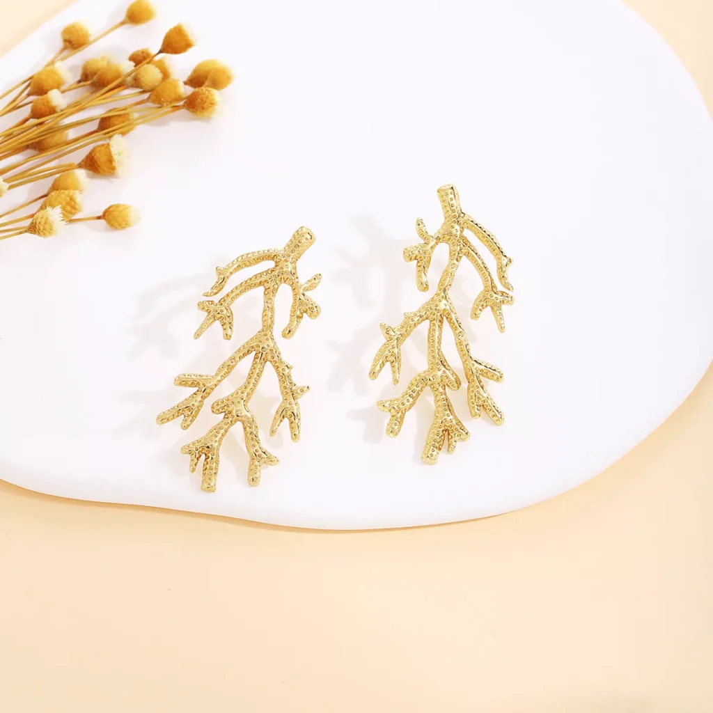 Gold coral branch design stainless steel earrings displayed on a white plate with dried flowers, elegant and nature-inspired wholesale jewelry.