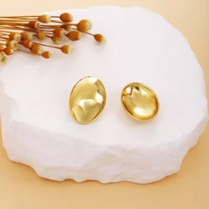 A pair of elegant oval-shaped gold-tone stainless steel stud earrings displayed on a white stone slab, with dried floral decor in the background, showcasing a smooth and polished surface for a luxurious look.