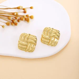 A pair of square-shaped gold-tone stainless steel earrings with an interwoven design, displayed on a white ceramic plate with dried floral accents, perfect for wholesale jewelry suppliers.