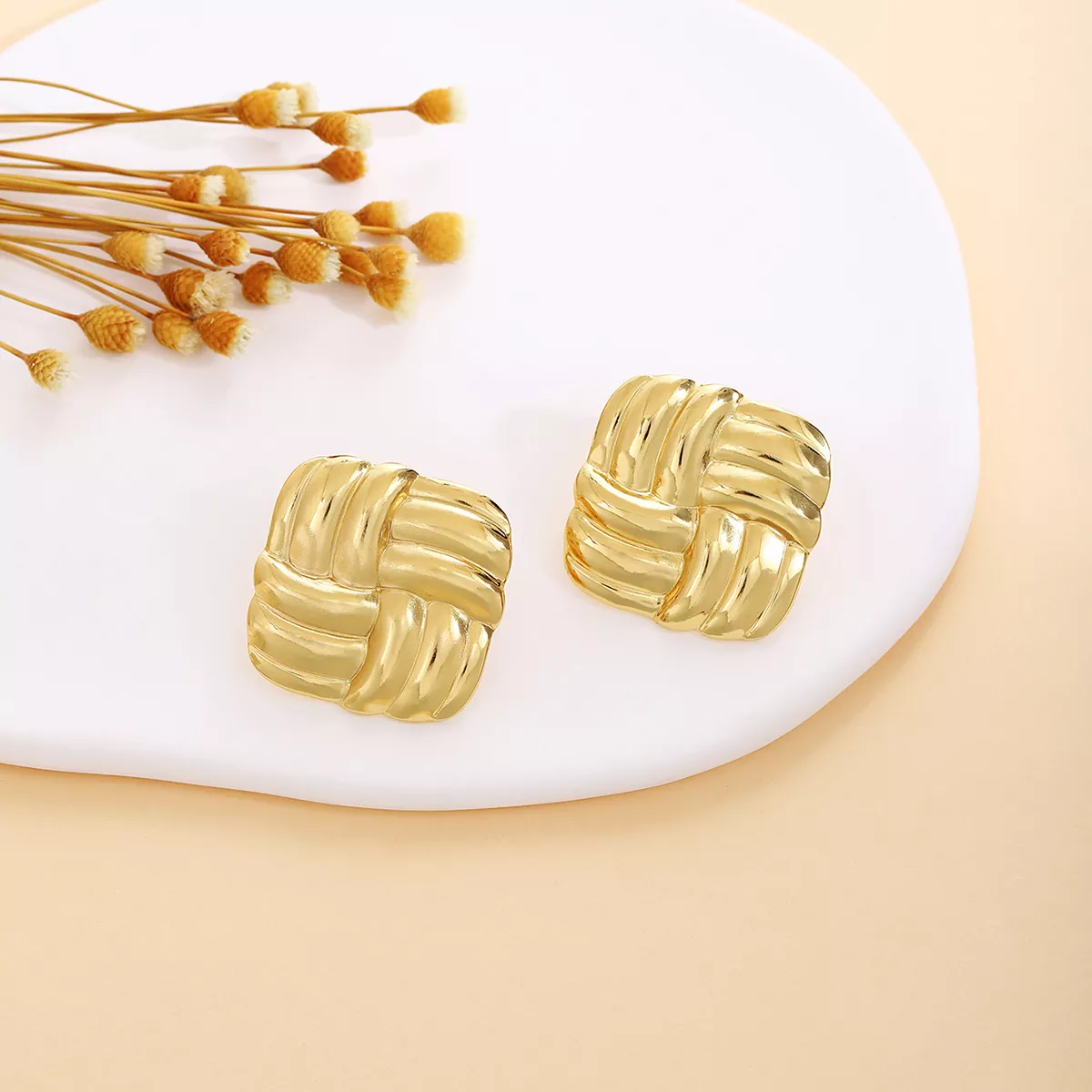 A pair of square-shaped gold-tone stainless steel earrings with an interwoven design, displayed on a white ceramic plate with dried floral accents, perfect for wholesale jewelry suppliers.